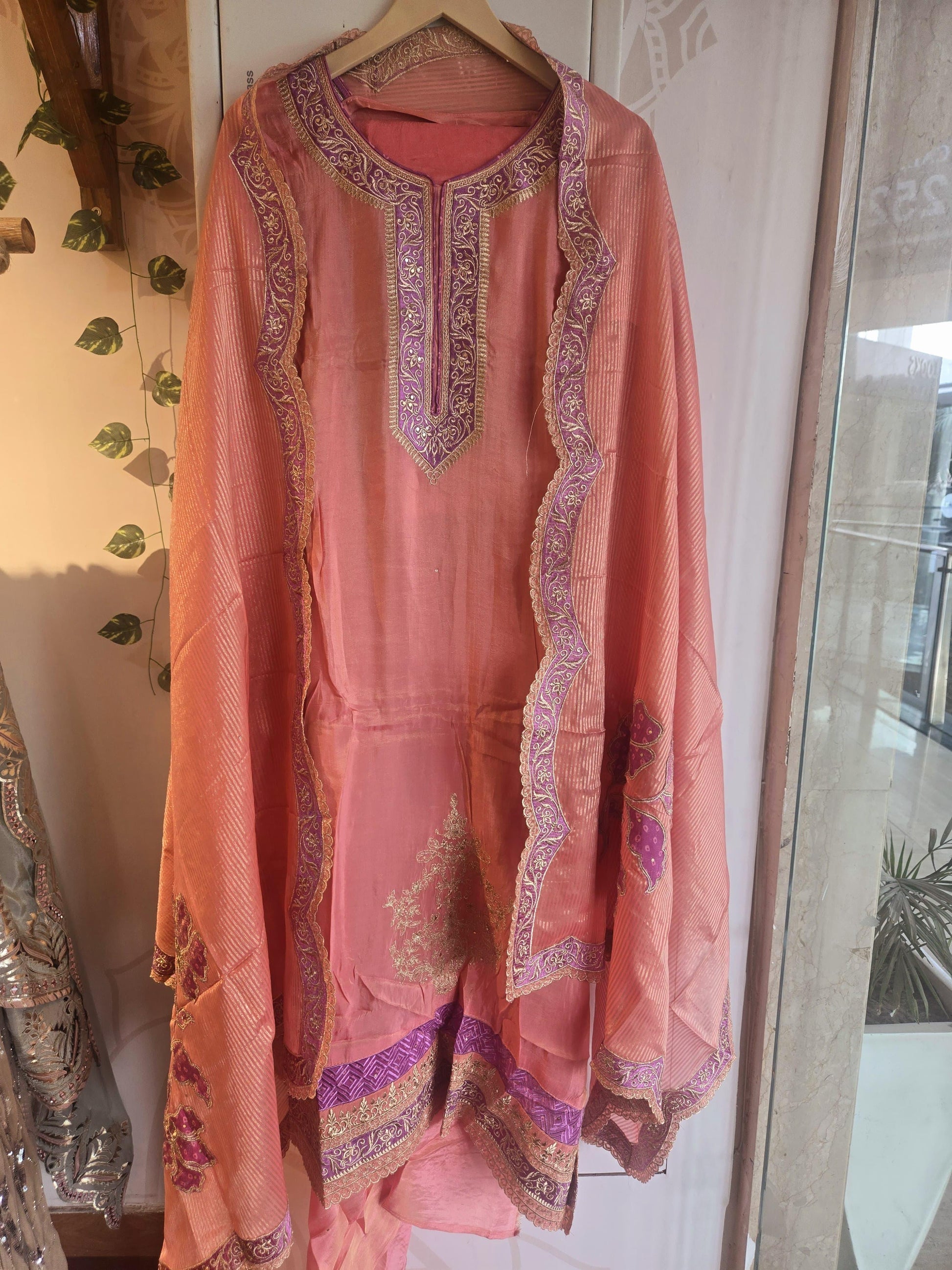 Peach shimmer unstitched suit with intricate embroidery hanging on a wooden hanger at Dharini's Store