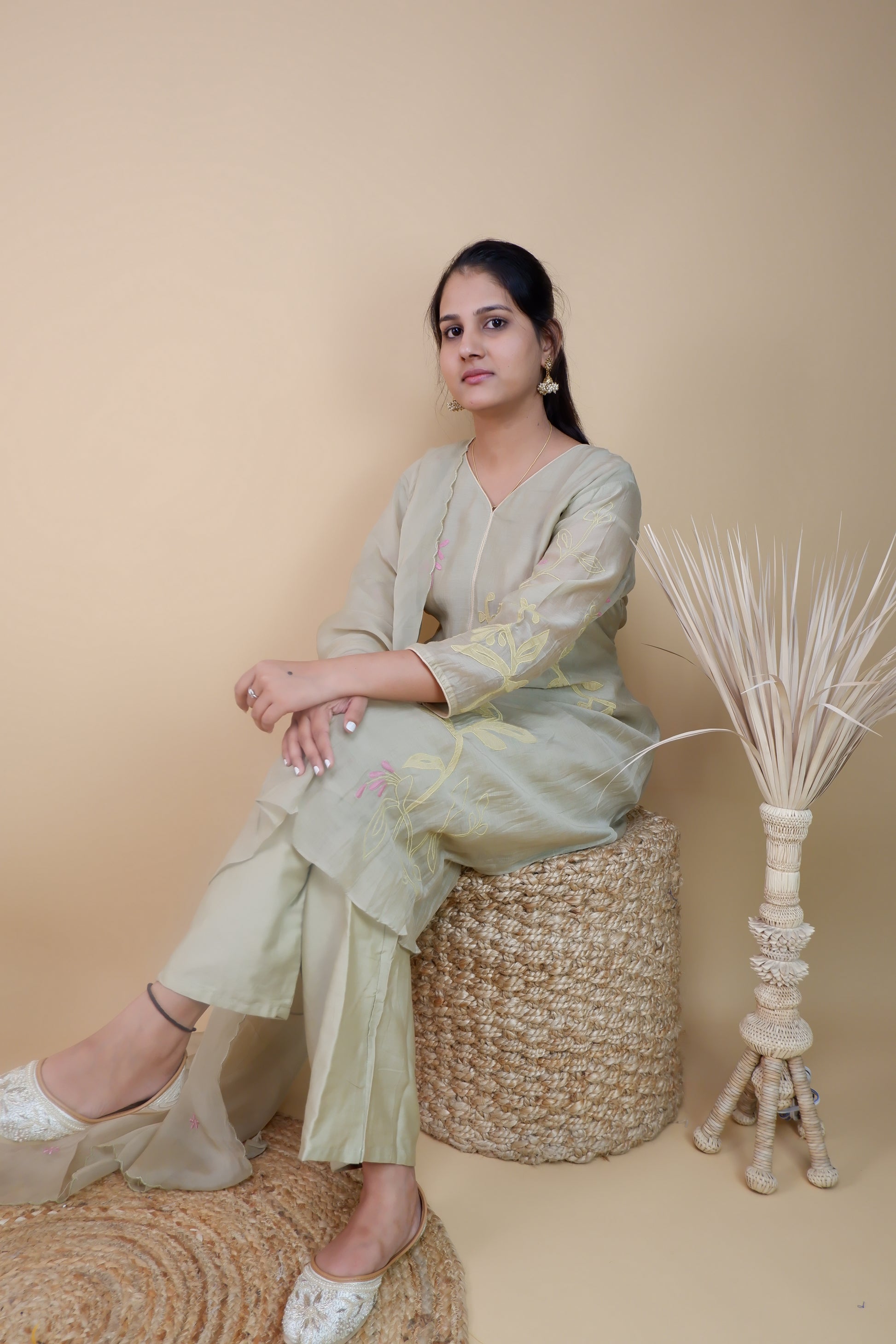Woman wearing olive chanderi embroidered suit sitting on woven stool in minimalist setting