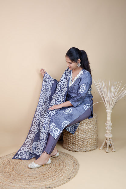Woman wearing grey and white cotton suit set sitting on a woven stool holding matching dupatta