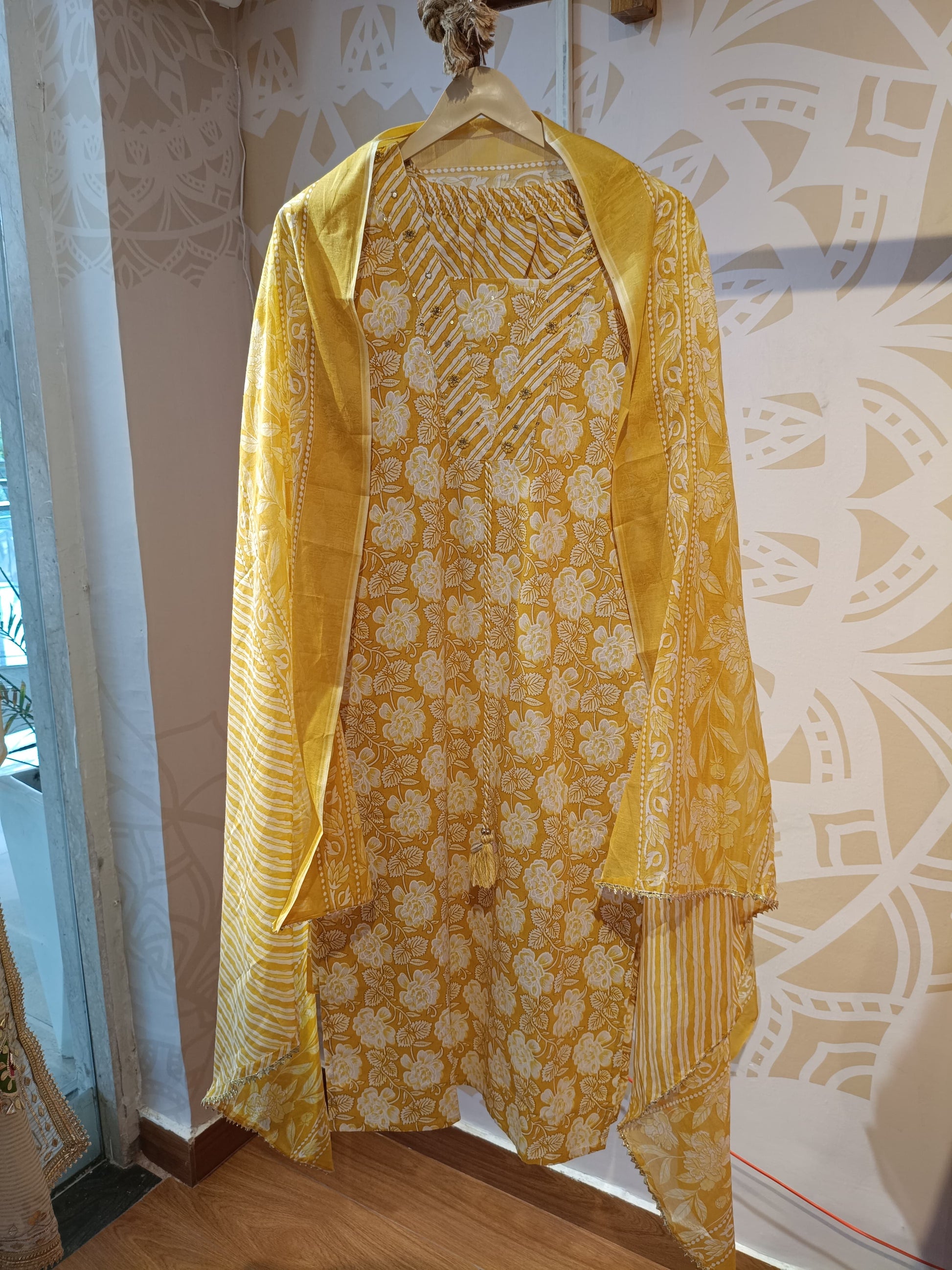 Yellow floral cotton suit with matching dupatta hanging on a wooden hanger at Dharini's Store